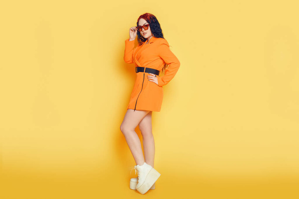 Beautiful girl in full growth on a yellow background. Brunette in orange dress and long hair posing. Woman with glasses, warm summer photo in the studio - Foto, Imagem