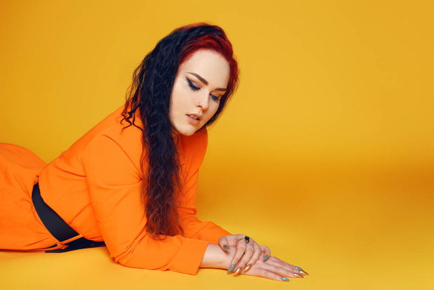 Nainen, jolla on pitkät kiharat hiukset, makaa lattialla keltaista taustaa vasten. Tyttö oranssissa mekossa, punaiset hiukset ja nuolet silmissä. Huippumuoti - Valokuva, kuva