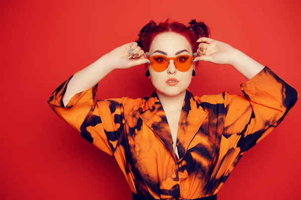 Cute girl on a red background. Red hair and tunnels in the ears, red dress and lips. Anime poses. A girl in orange sunglasses and two buns on her head looks at the camera. - Photo, Image