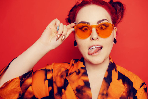 A woman in red sunglasses and two buns on her head looks at her nose, grimaces, pierced tongue.Cute girl on a red background. Red hair and tunnels in the ears, red dress and lips.  - Foto, afbeelding