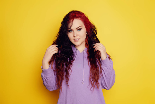 Femme dans un pull violet sur un fond jaune posant. Une belle brune aux longs cheveux bouclés et aux racines rouges. Eyeliner et maquillage tendance. Modèle regardant la caméra - Photo, image