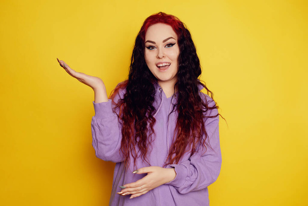 Woman in a purple sweater on a yellow background posing. A beautiful brunette with long curly hair and red roots. The model looks at the camera and points to an empty space, holds the product - Fotoğraf, Görsel