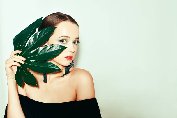 Sexy beauty brunette model with green and blue make up and green leaves. Copy space. Close up, selective focus. Girl with tropical leaf covers a part of her face. Fashion, cosmetics, accessories. - Valokuva, kuva