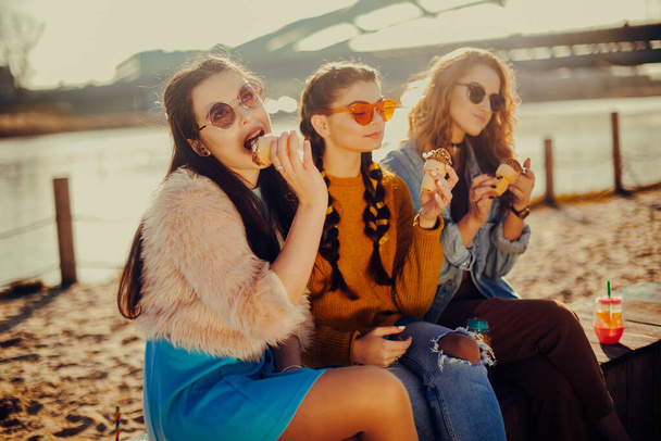 Trois filles s'assoient sur la plage, près de la rivière, parlent, bavardent, plaisantent, rient, sourient à la journée des femmes. Par une chaude journée, les filles mangent de la glace. Modèle beauté portant des lunettes de soleil le jour d'été. Fille avec hairslyle - Photo, image