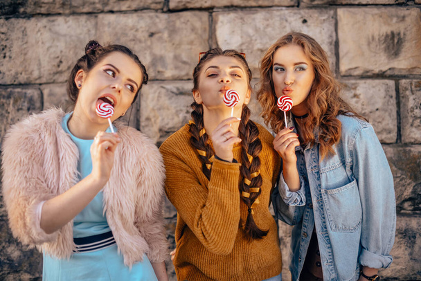 Beautiful teenage girlfriends in trendy dresses and glasses walk in the summer. Sunny day, summer mood, fun and happiness. Three girls are smiling and eating a lollipop. - Photo, Image
