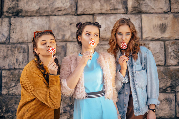 Beautiful teenage girlfriends in trendy dresses and glasses walk in the summer. Sunny day, summer mood, fun and happiness. Three girls are smiling and eating a lollipop. - Фото, изображение