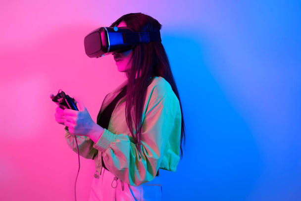 Γέλιο χαμογελαστό κορίτσι έκπληξη σε νέον με γυαλιά VR κρατά ένα gamepad στα χέρια της και παίζει παιχνίδια. Εικονικός κόσμος, τεχνολογία. - Φωτογραφία, εικόνα