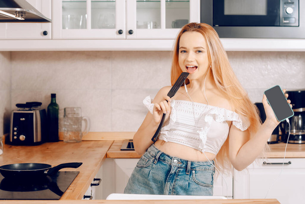  Frau singt und tanzt in der Küche mit Smartphone, Musik. Ein Mädchen mit Kopfhörer und Telefon steht in der Küche und bereitet hinter einem Induktionsherd Essen zu - Foto, Bild