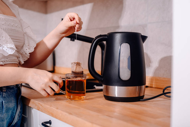 Egy fekete vízforraló van a konyhaasztalon. Egy nő keze teáskannát fog és teát főz. Tea zsák egy bögrében vízzel, tealevelek. - Fotó, kép