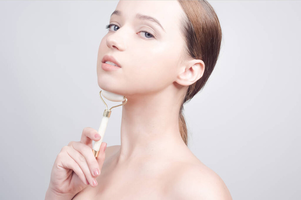 Beauty face care. Woman doing face massage with white jade facial rollers for spa skin care treatment at home. Girl model using natural massager tool portrait on white  - 写真・画像