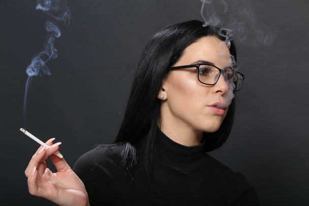 Houkutteleva eurooppalainen tyttö, jolla on mustat hiukset ja lasit ja joka poseeraa studiossa ja polttaa sikaria eristetyllä taustalla. Tyyli, trendit, tupakointi käsite - Valokuva, kuva