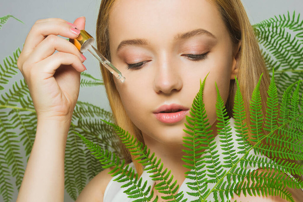 Lány a háttérben a trópusi levelek természetes kozmetikumok és gyógyszerek. A modell egy palackot és olajat tart az arcon, és a bőrre alkalmazza. Palack pipettával. - Fotó, kép