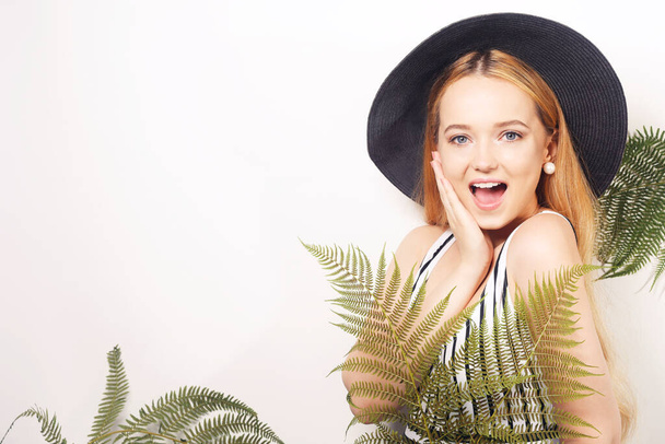 Girl in tropical leaves in a swimsuit in a big black hat smiles. Space for text, empty space point hand. Model summer photo. Big earrings - Photo, Image