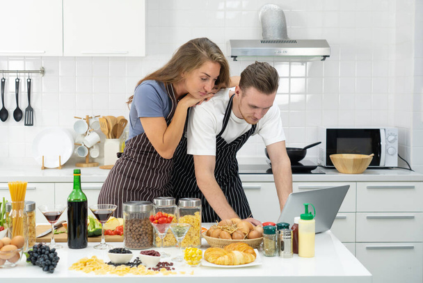young caucasian couple reading instruction of new cuisine in computer before cooking food in kitchen - Photo, Image