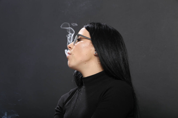 Atrakcyjna Europejka z czarnymi włosami i okularami, pozująca w studio i paląca cygaro na odosobnionym tle. Styl, trendy, koncepcja palenia - Zdjęcie, obraz