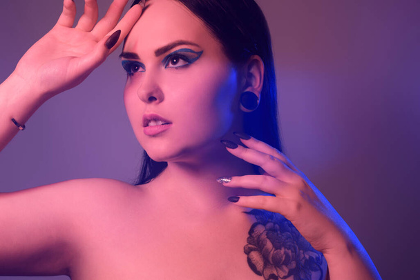 Brunette girl with a flower tattoo on her shoulder and tunnels in her ears in beautiful blue light. Woman with purple make-up, fashionable blue arrows over the eyelid. - Foto, Imagem
