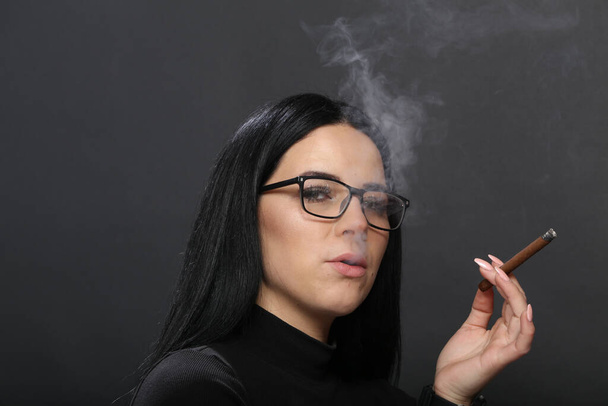 スタジオでポーズをとったり、孤立した背景で葉巻を吸っている黒い髪と眼鏡を持つ魅力的なヨーロッパの女の子。スタイル、トレンド、喫煙コンセプト - 写真・画像