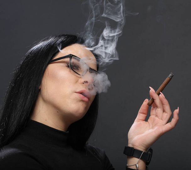 Приваблива дівчина з чорним волоссям і окулярами, що сидять у студії і курять сигару на ізольованому фоні. Стиль, тенденції, поняття куріння - Фото, зображення