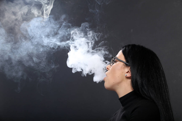 Ελκυστική Ευρωπαία με μαύρα μαλλιά και γυαλιά που ποζάρει στο στούντιο και καπνίζει πούρο σε απομονωμένο φόντο. Στυλ, τάσεις, έννοια του καπνίσματος - Φωτογραφία, εικόνα
