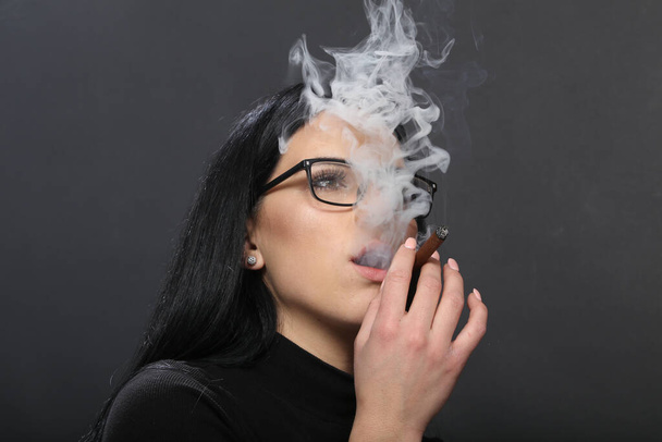 Attractive european girl with black hair and glasses posing in studio and smoking a cigar on isolated background. Style, trends, smoking concept - Foto, afbeelding