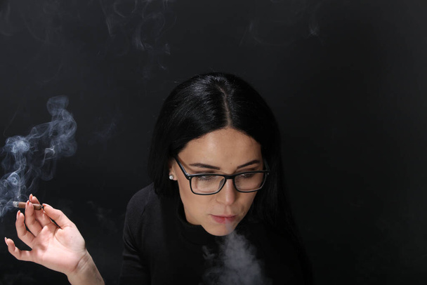 Attractive european girl with black hair and glasses posing in studio and smoking a cigar on isolated background. Style, trends, smoking concept - Photo, Image