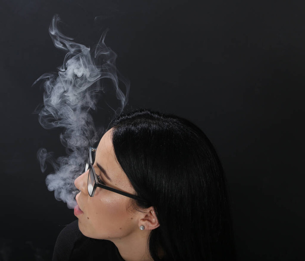 Atractiva chica europea con el pelo negro y gafas posando en el estudio y fumando un cigarro sobre un fondo aislado. Estilo, tendencias, concepto de fumar - Foto, imagen