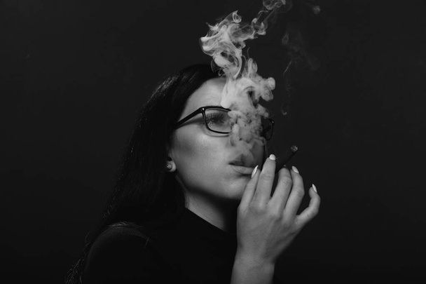 Siyah saçlı, gözlüklü, stüdyoda poz veren ve arka planda puro içen çekici Avrupalı bir kız. Tarz, trendler, sigara konsepti - Fotoğraf, Görsel