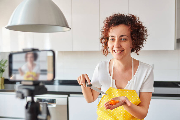 La mujer youtuber se graba con su smartphone en su cocina mientras prepara una receta de ensalada, hay luz natural y lleva un delantal amarillo y una camiseta blanca, es caucásica - Foto, imagen