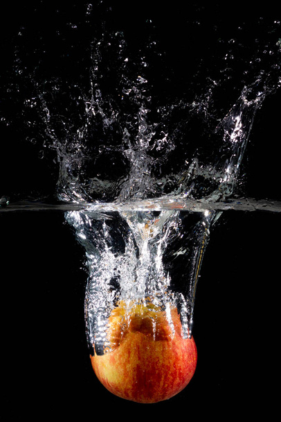 A vertical shot of a red apple in water with splashes on a dark isolated background - Fotografie, Obrázek