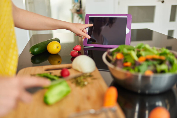 Joven mujer caucásica prepara una ensalada en la cocina de su casa mientras mira su tableta digital, es de día y hay luz natural, lleva un delantal amarillo - Foto, imagen