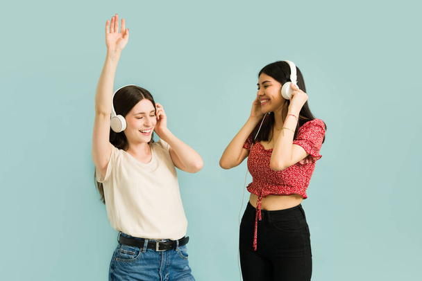 Kaunis monirotuinen nuoret naiset kuuntelevat onnellista musiikkia kuulokkeet ja alkaa tanssia sinistä taustaa vasten - Valokuva, kuva