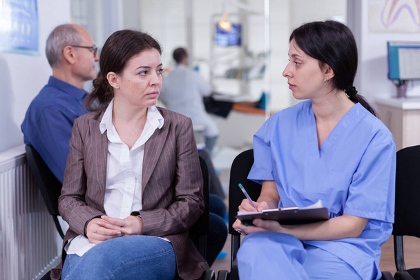 Enfermera tomando notas en portapapeles discutiendo con paciente en clínica dental - Foto, Imagen