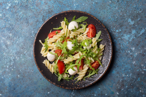 Top view of tasty vegetarian pasta with cherry tomatoes, soft cheese mozzarella and green arugula leaves on blue concrete background - Fotografie, Obrázek