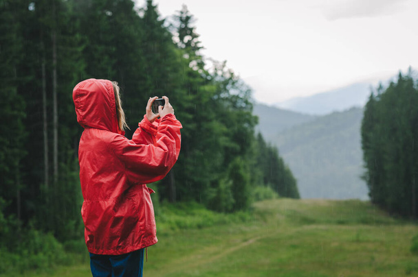 Πεζοπόρος γυναίκα με κόκκινο αδιάβροχο στέκεται στο παρασκήνιο του όμορφου τοπίου στο βουνό σε βροχερές καιρικές συνθήκες και παίρνει φωτογραφία στο smartphone. Κορίτσι λήψη φωτογραφιών του τοπίου στα βουνά κατά τη διάρκεια της βροχής στην πεζοπορία. - Φωτογραφία, εικόνα
