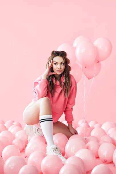 Bored fashionable woman sitting on floor with pink air balloons on pink pastel background - Photo, Image