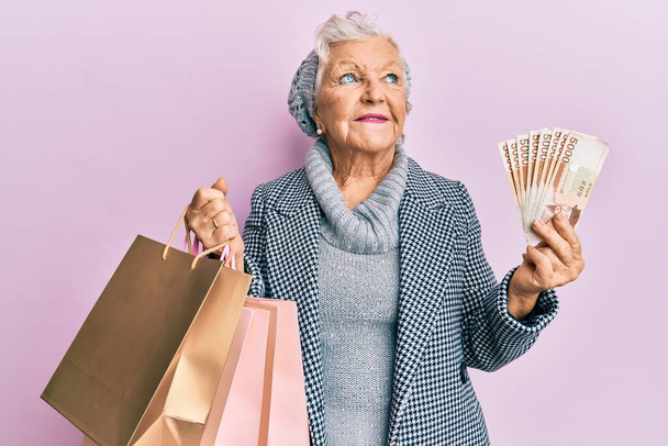 Ältere grauhaarige Frau mit Einkaufstaschen und südkoreanischen Geldscheinen lächelt zur Seite und starrt nachdenklich weg.  - Foto, Bild