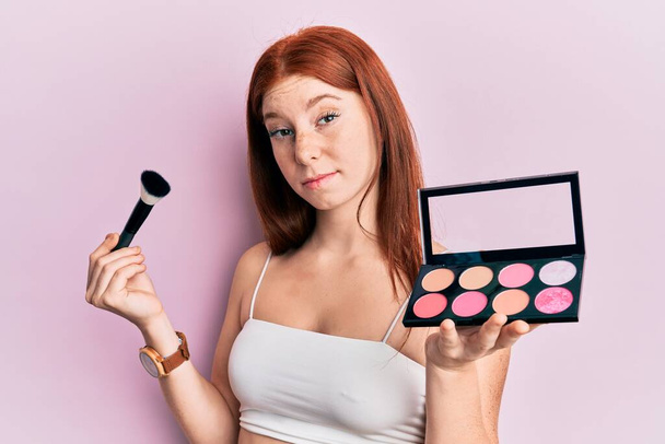 Young red head girl holding makeup brush and blush palette skeptic and nervous, frowning upset because of problem. negative person.  - Photo, Image