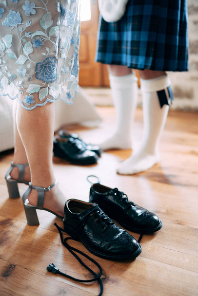 Vorbereitungen für eine schottische Hochzeit. Mann im Kilt und Sporn steht neben Frau im Rock und Stöckelschuhen. Auf dem Boden liegen Schuhe mit langen Schnürsenkeln. Seitenansicht - Foto, Bild