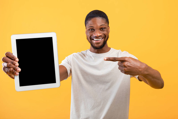 Positivo ragazzo afroamericano che punta al tablet con schermo nero bianco su sfondo giallo, spazio per il mockup - Foto, immagini