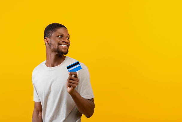 Emocionado hombre afroamericano con tarjeta de crédito y mirando el espacio vacío en el fondo del estudio amarillo - Foto, Imagen