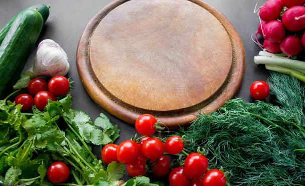 Kasa tuoreita, kypsiä vihanneksia: retiisit, tomaatit, kurkut, yrtit ja valkosipuli on järjestetty pyöreän puisen leikkuulaudan ympärille. Kopioi tila. - Valokuva, kuva