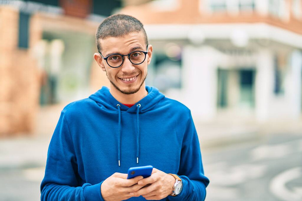 Молодий іспаномовний чоловік посміхається щасливим, використовуючи смартфон у місті
. - Фото, зображення