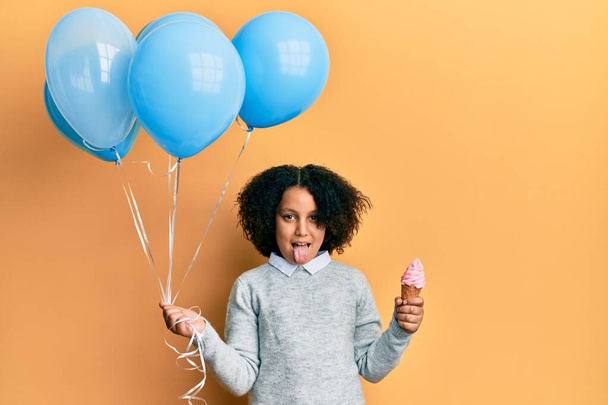 Giovane bambina con i capelli afro in possesso di gelato e palloncini blu attaccare la lingua fuori felice con espressione divertente.  - Foto, immagini