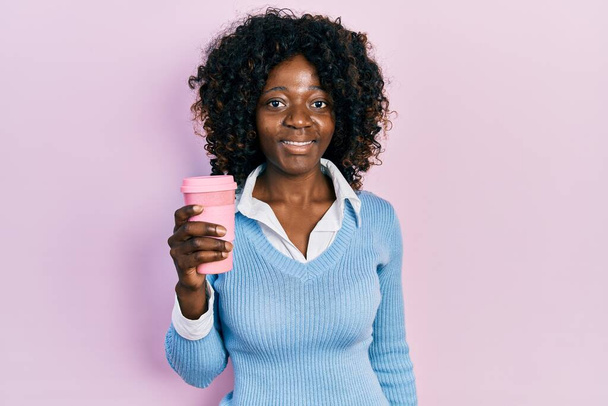 Młoda Afroamerykanka pijąca kawę zabiera ją wyglądając pozytywnie i szczęśliwie stojąc i uśmiechając się z ufnym uśmiechem pokazującym zęby  - Zdjęcie, obraz