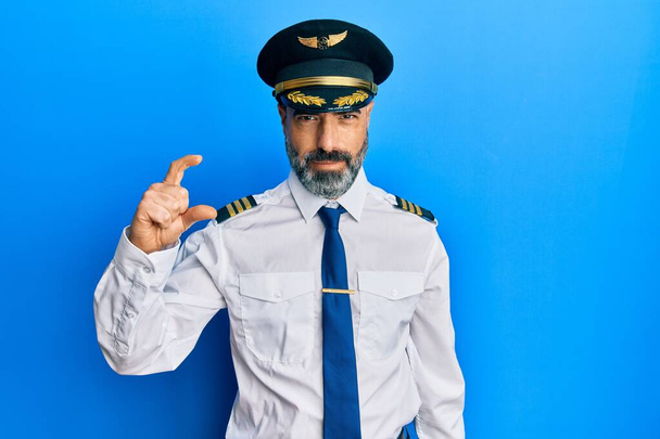 Middle age man with beard and grey hair wearing airplane pilot uniform smiling and confident gesturing with hand doing small size sign with fingers looking and the camera. measure concept.  - Photo, Image