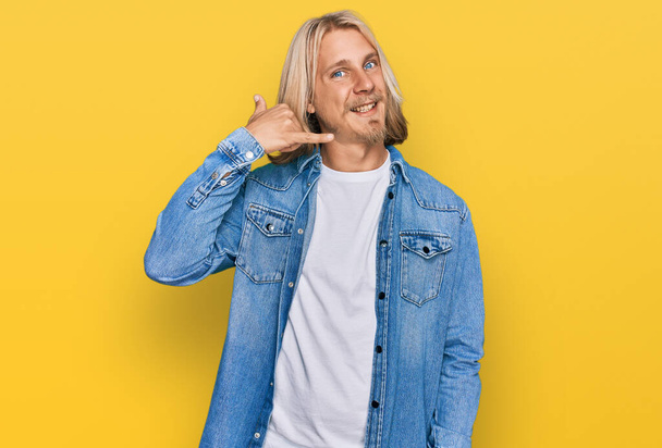 Homme caucasien aux cheveux longs blonds portant une veste en denim décontractée souriant faisant geste téléphonique avec la main et les doigts comme parler au téléphone. communiquer les concepts.  - Photo, image