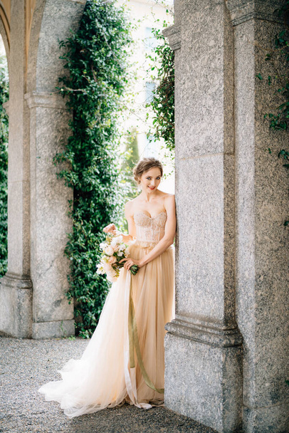 Улыбающаяся невеста в платье с букетом цветов стоит у арки старинной виллы. Озеро Комо, Италия - Фото, изображение