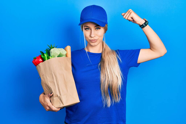 Молодая белая женщина в форме курьера с продуктами из супермаркета сильный человек показывает мышцы рук, уверенный и гордый властью  - Фото, изображение
