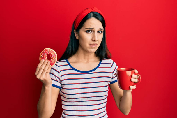 若いヒスパニック系の女性がドーナツを食べ、コーヒーを飲みながら混乱した表情。疑わしい概念.  - 写真・画像