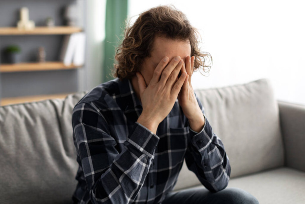 Δυστυχισμένος τύπος που καλύπτει το πρόσωπο που υποφέρει από κατάθλιψη κάθεται στο σπίτι - Φωτογραφία, εικόνα